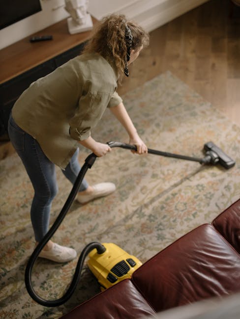 Close-up of a modern, portable industrial vacuum cleaner with a transparent dust container filled with debris, connected to a flexible suction hose. The vacuum is positioned on a pink carpeted floor i
